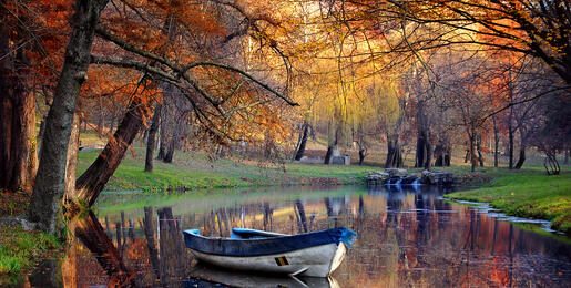 Autumn scene of a boat moored at the side of a lazy river under trees covered with orange and red autumn leaves.................
